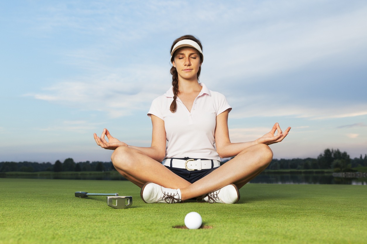 A female golfer sitting in meditation on the grass with club and golfball close to the whole